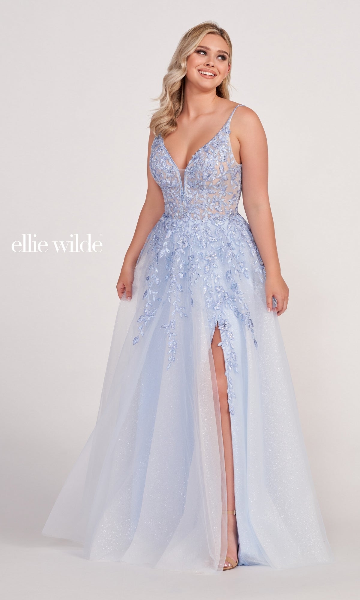 A woman wears the Glitter-Tulle Long Ellie Wilde Prom Dress EW34089, featuring a light blue sleeveless design with embroidered lace, floral appliqué, deep V-neckline, and high slit against a plain backdrop.